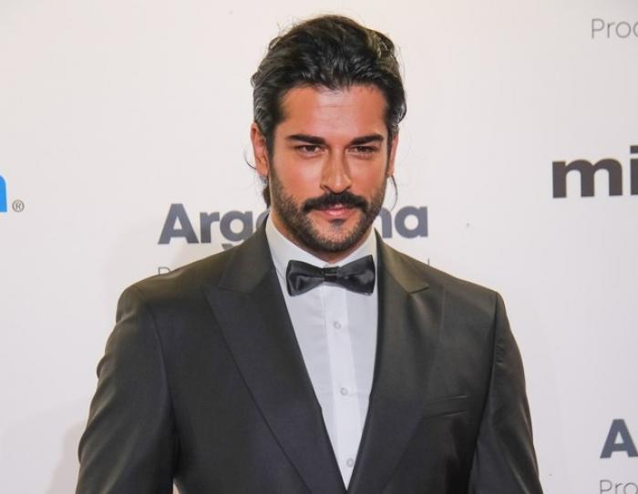 Burak Özçivit poartă o cămașă albă, costum negru și papion. Actorul zâmbește, are mustață și părul negru