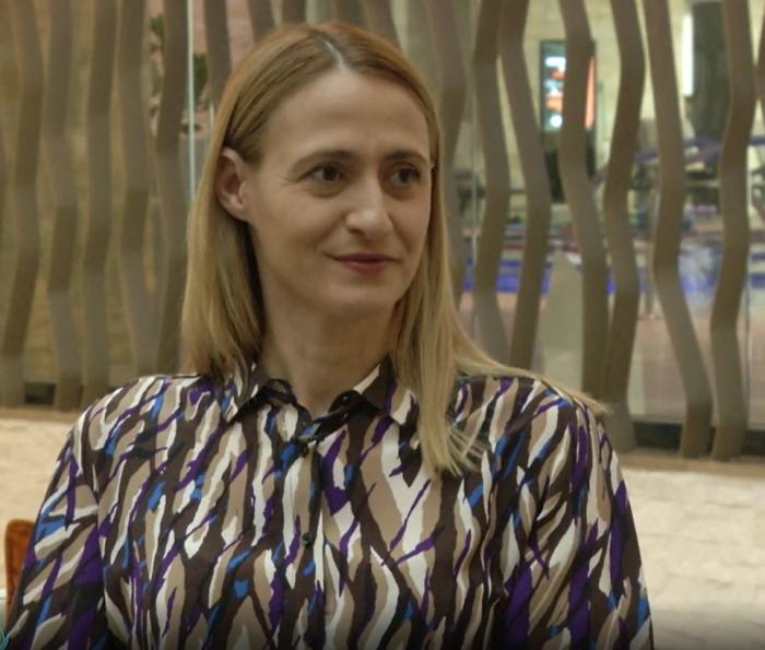 Camelia Potec poartă părul lung, desprins și blond. Campioana are o bluză cu imprimeu și zâmbește cu gura închisă