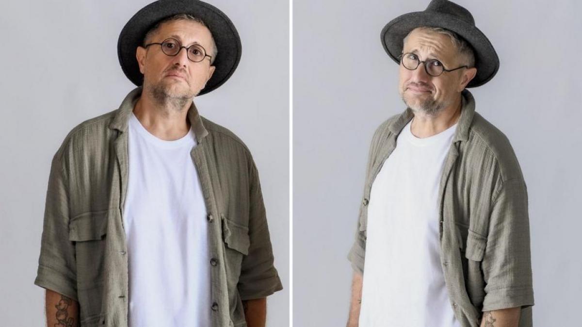 Bogdan Talașman poartă un tricou alb, o cămașă kaki, pălărie și ochelari de vedere