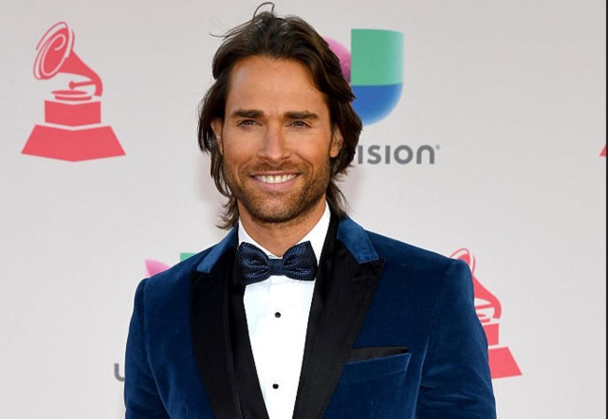 Sebastián Rulli poartă un costum închis la culoare, cămașă albă și papion. Actorul are părul lung