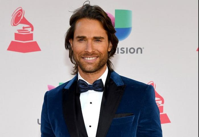 Sebastián Rulli poartă un costum închis la culoare, cămașă albă și papion. Actorul are părul lung