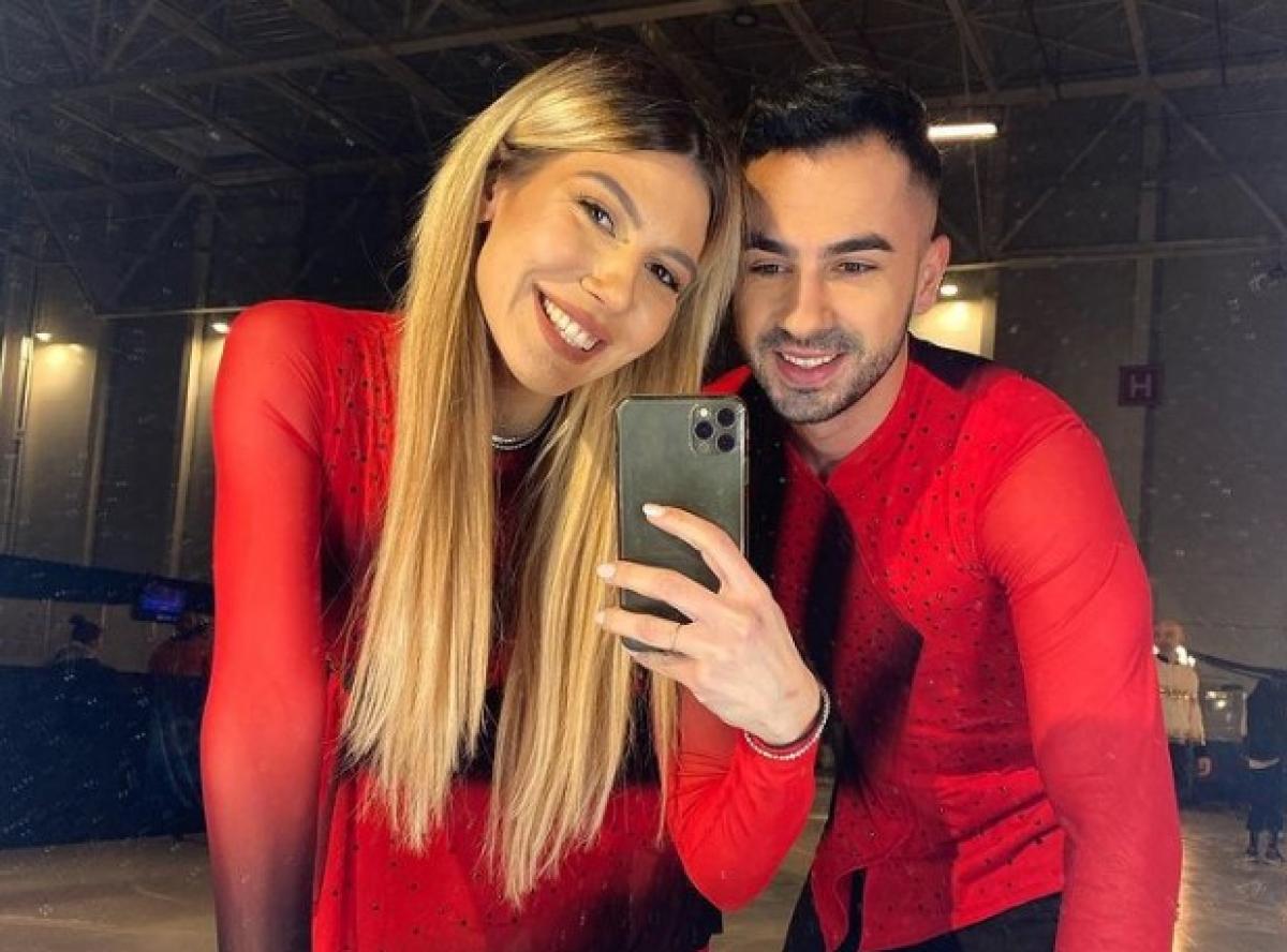 Sore poartă un costum roșu de gală, are părul desprins, lung și stă alături de Grațiano Dinu