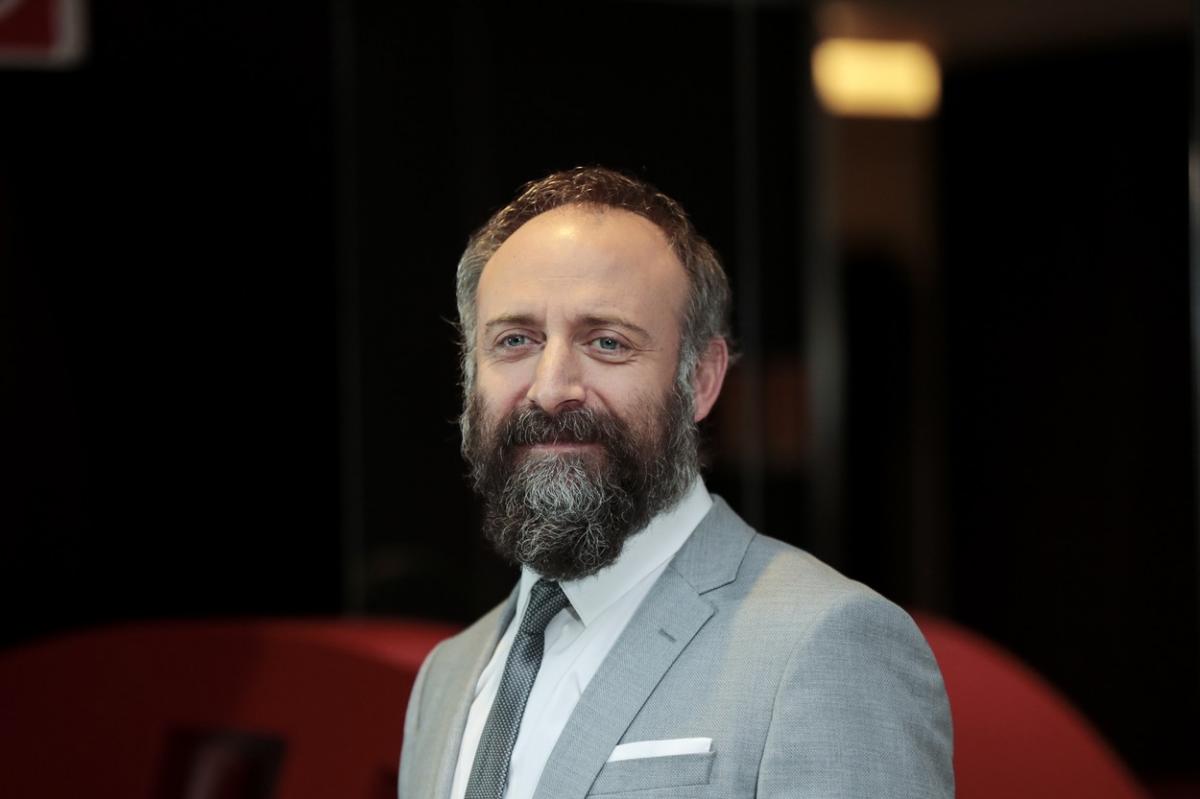 Halit Ergenç poartă cămașă albă, costum gri și cravată. Actorul zâmbește și are barbă și mustață