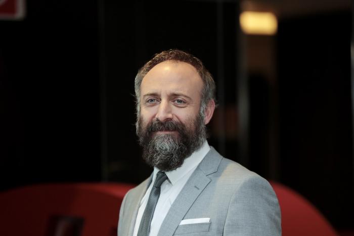 Halit Ergenç poartă cămașă albă, costum gri și cravată. Actorul zâmbește și are barbă și mustață