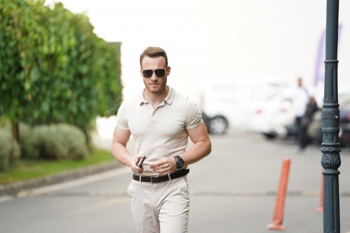 Kerem Bürsin poartă pantaloni deschiși la culoare și tricou asemănător. Are ochelari negri și privește în depărtare