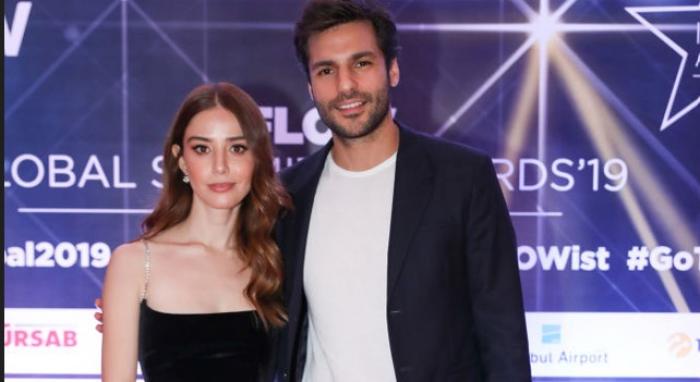 Özge Gürel și Serkan Çayoğlu stau unul lângă celălalt și zâmbesc. Actrița este îmbrăcată într-o rochie neagră fără mâneci, iar partenerul ei poartă un costum negru și o cămașă albă