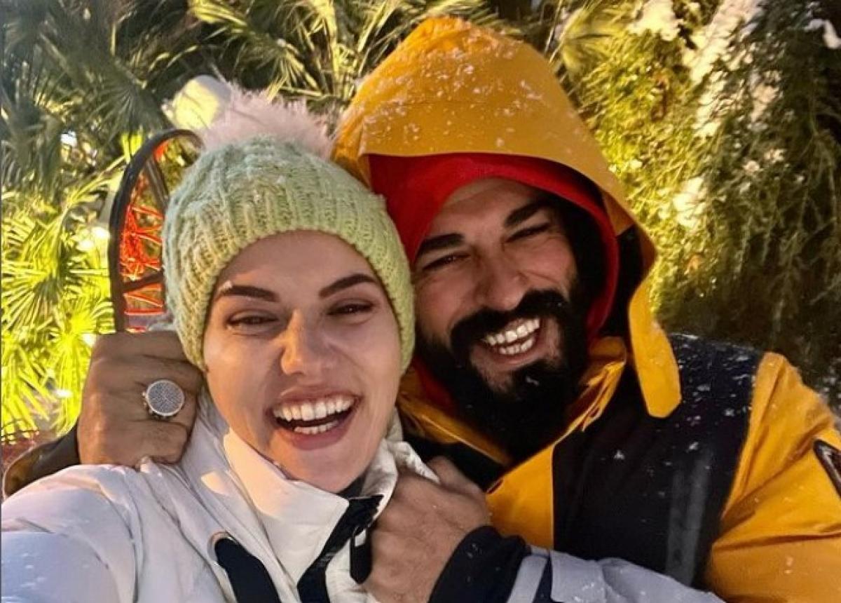 Fahriye Evcen poartă o geacă albă și zâmbește, stă alături de soțul ei, Burak Özçivit