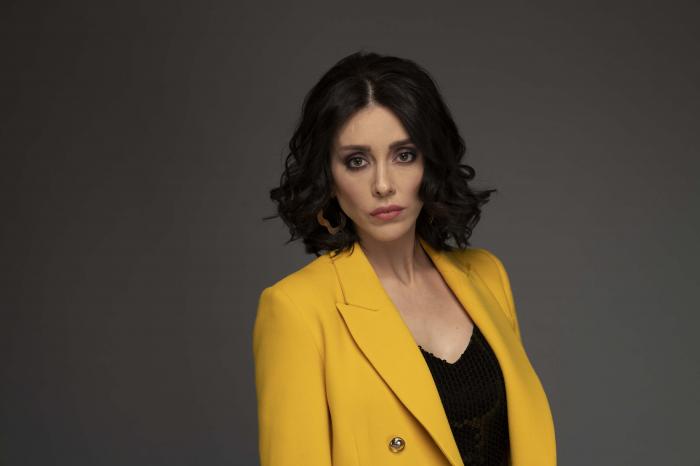 Elif Erol poartă o bluză neagră și un sacou galben și are părul desprins