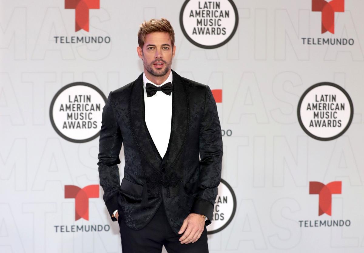 William Levy poartă un costum negru, cămașă albă și papion pe covorul roșu. Actorul își ține mâna dreaptă în buzunar