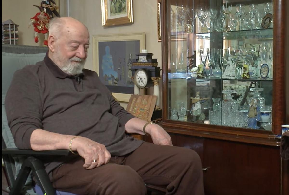 Constantin Dinulescu se află în casa lui, așezat pe un fotoliu cu mânere. Își ține mâinile sprijinite și are un zâmbet larg pe față