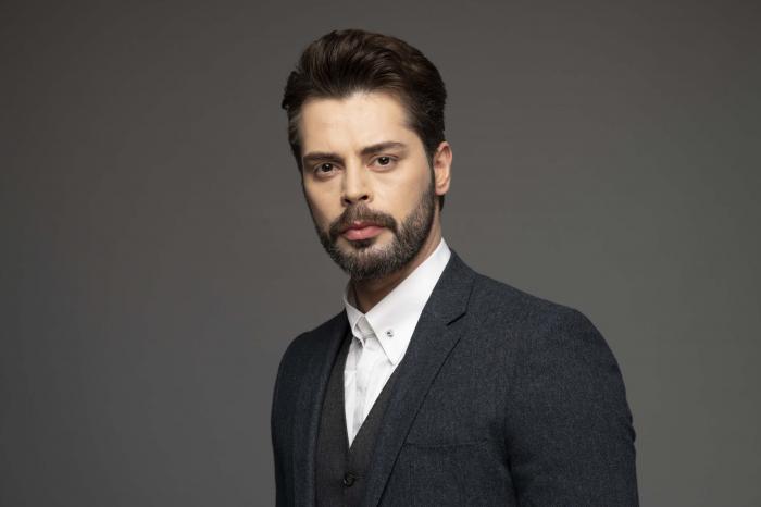 Ali Yasin Özegemen poartă o cămașă albă și un sacou negru. Actorul are barbă, mustață și privește serios către cameră