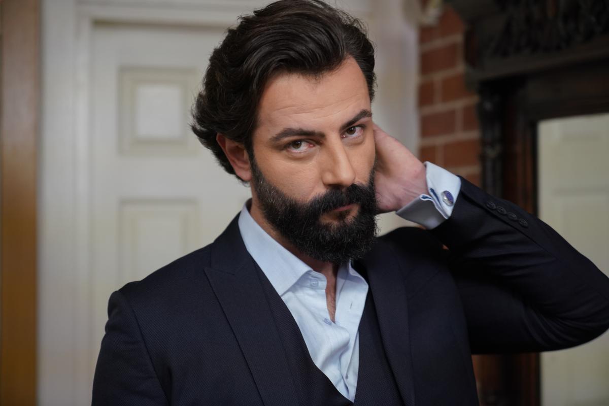 Gökberk Demirci poartă o cămașă deschisă la culoare și un sacou negru și îți ține mâna stângă în păr