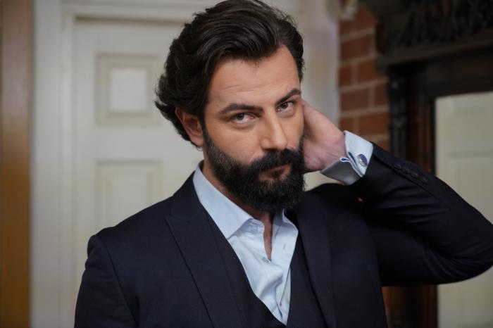 Gökberk Demirci poartă o cămașă deschisă la culoare și un sacou negru și îți ține mâna stângă în păr