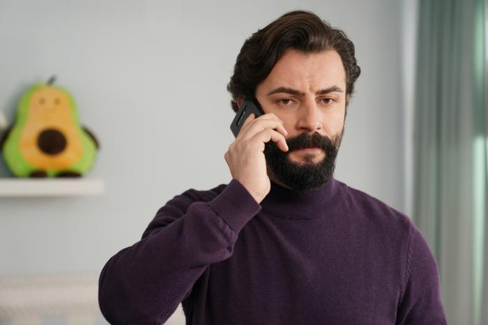 Gökberk Demirci poartă un pulover maro închis și ține un telefon la ureche cu mâna dreaptă