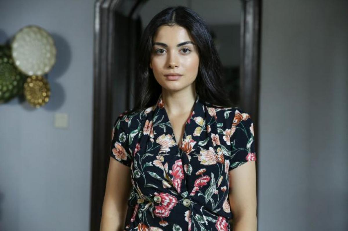 Özge Yağız poartă o rochie cu flori cu mâneci scurte, are părul lung și desprins și se uită în depărtare