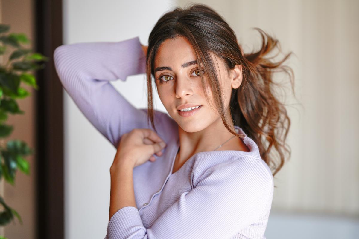 Sıla Türkoğlu poartă o bluză deschisă la culoare și îți ține părul prins cu mâna dreaptă