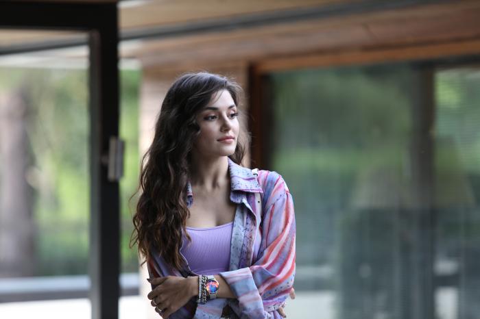 Hande Erçel poartă o bluză mov cu mâneci scurte și se uită în depărtare