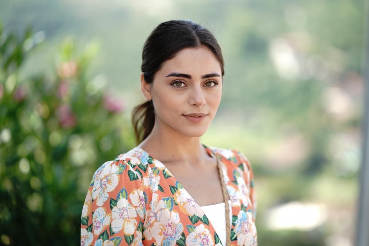 Sıla Türkoğlu poartă un tricou alb și o rochie cu flori. Actrița are părul prins într-o coadă joasă, la spate