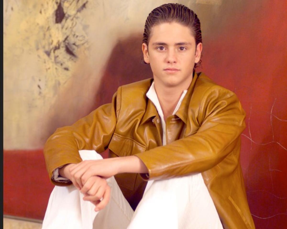 Christopher Uckermann poartă pantaloni albi lungi și o geacă de piele maro. Stă pe jos cu mâna dreaptă așezată pe genunchiul drept