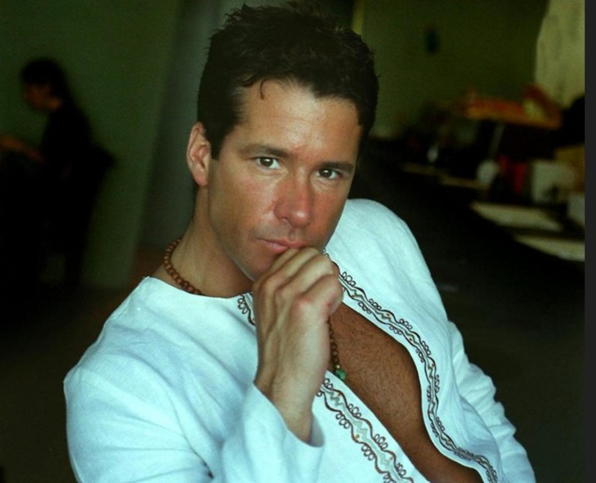 Fernando Carillo poartă o bluză deschisă și își ține mâna dreaptă în dreptul gurii