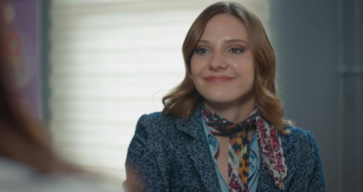 Alina Boz poartă un costum, o eșarfă la gât, are părul desprins și zâmbește