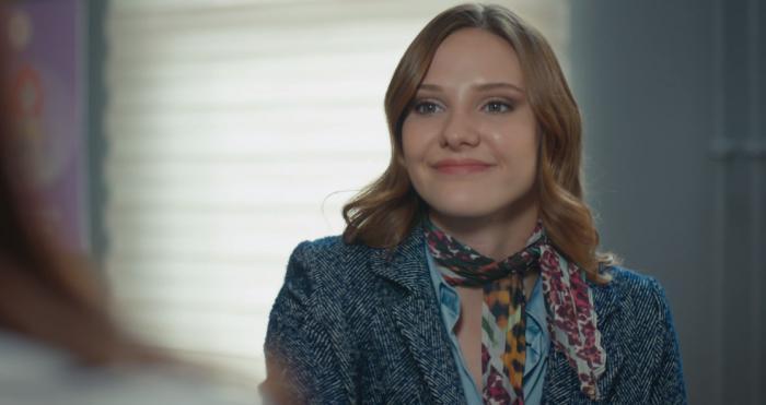 Alina Boz poartă un costum, o eșarfă la gât, are părul desprins și zâmbește