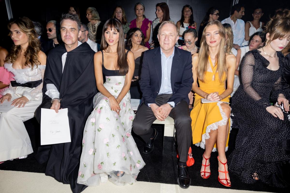 Eiza Gonzalez apare îmbrăcată într-o fustă lungă albă cu imprimeu floral și un top negru și stă alături de Robbie Williams în cadrul unei prezentări de modă