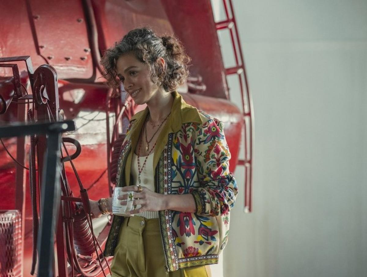 Özge Özpirinççi poartă pantaloni verde muștar, are părul scurt și ține în mâna stângă o cană