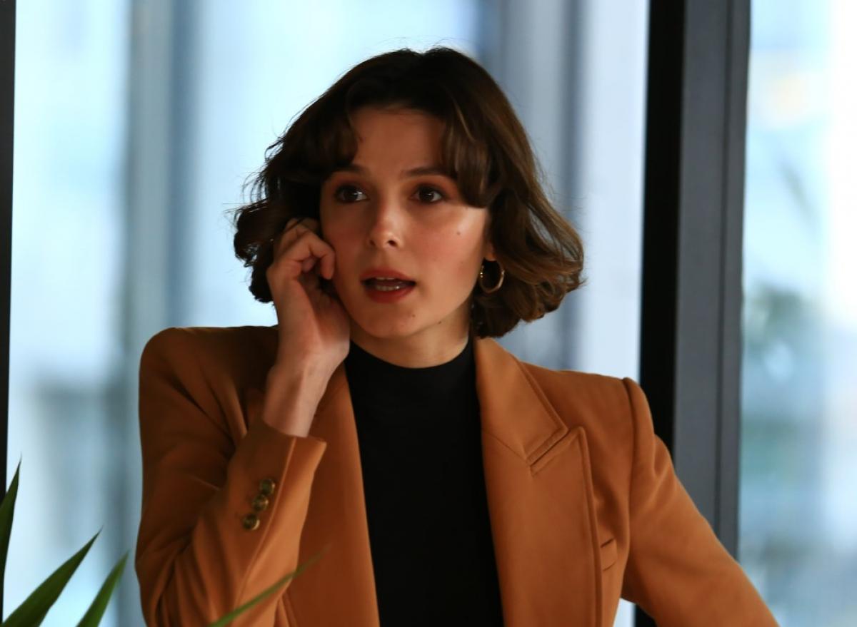 Büşra Develi poartă o bluză neagră și un sacou maro și ține telefonul la ureche cu mâna dreaptă