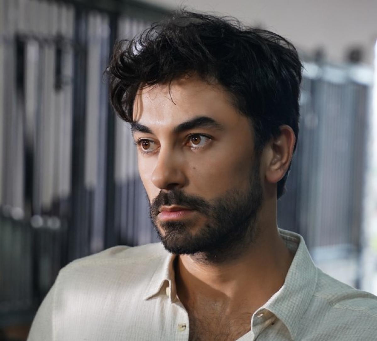 Gökhan Alkan poartă o bluză albă, are barbă și mustață și se uită în depărtare