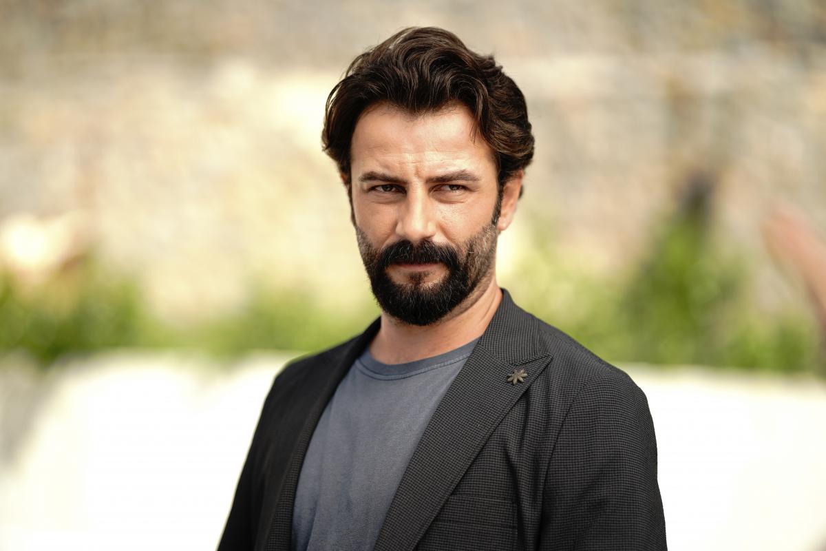 Gökberk Demirci poartă un tricou închis la culoare și un sacou maro