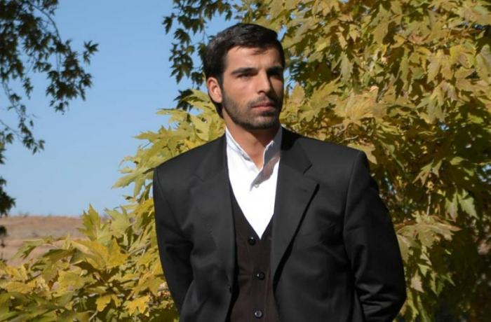 Mehmet Akif Alakurt poartă o cămașă albă și un sacou negru și își ține mâinile la spate