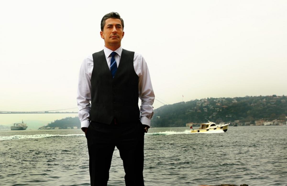 Erkan Petekkaya poartă o cămașă deschisă la culoare și un sacou negru și își ține mâinile în buzunare