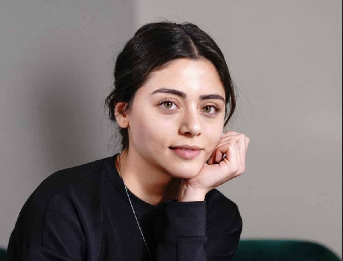 Sıla Türkoğlu poartă o bluză neagră cu mâneci lungi, zâmbește și își ține mâna stângă în dreptul feței