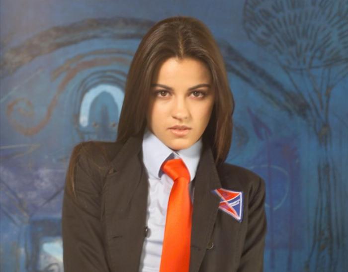 Maite Perroni poartă o cămașă albastră și un sacou negru și o cravată roșie și are părul lung desprins, coafat pe spate