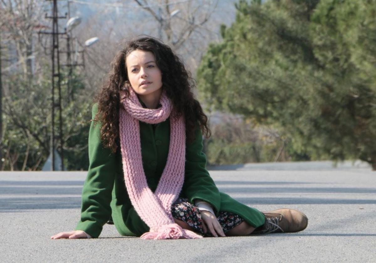 Gizem Arıkan poartă un palton verde și un fular roz și stă pe jos, în stradă