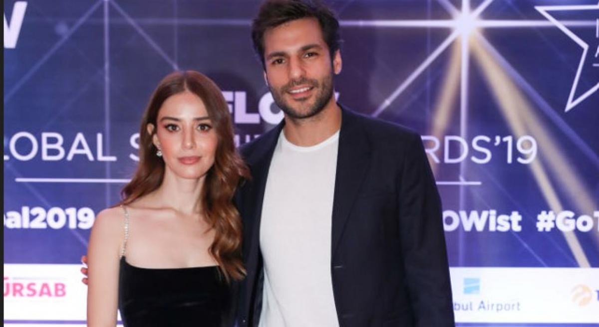 Özge Gürel și Serkan Çayoğlu stau unul lângă celălalt și zâmbesc. Actrița este îmbrăcată într-o rochie neagră fără mâneci, iar partenerul ei poartă un costum negru și o cămașă albă