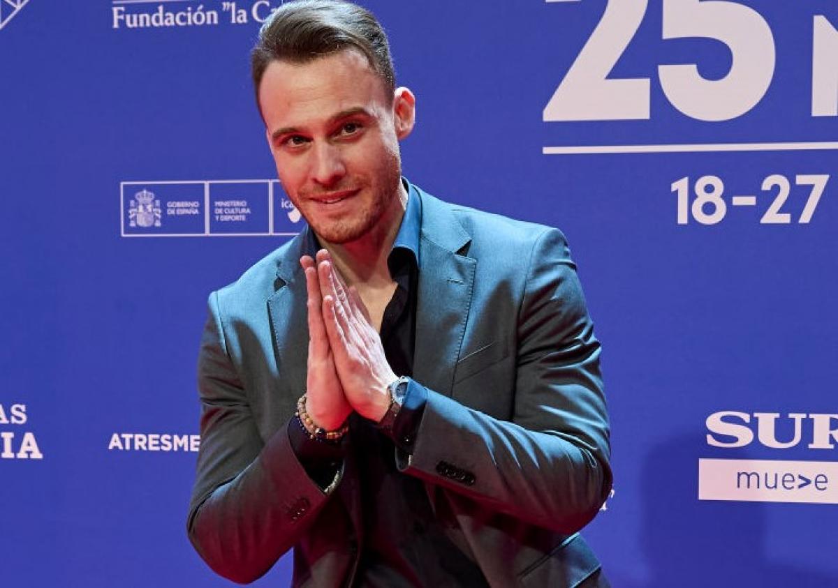 Kerem Bürsin poartă un costum închis la culoare și își ține mâinile împreunate