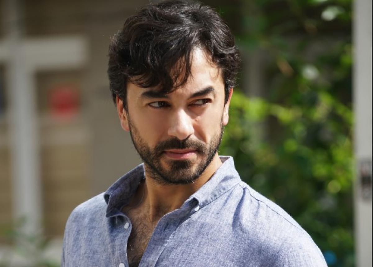 Gökhan Alkan poartă o cămașă ablastră deschisă la gât și privește curios