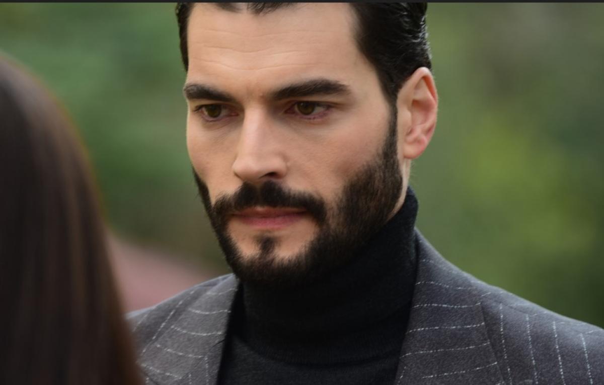 Akın Akınözü poartă o maletă neagră și un sacou gri