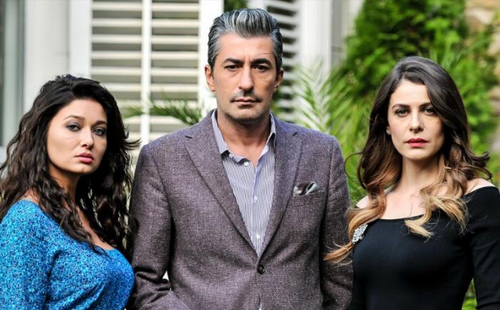 Erkan Petekkaya poartă un costum gri și stă între două actrițe