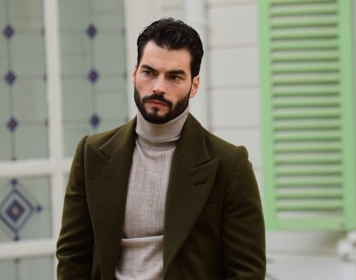 Akın Akınözü poartă o maletă maro și un sacou închis la culoare