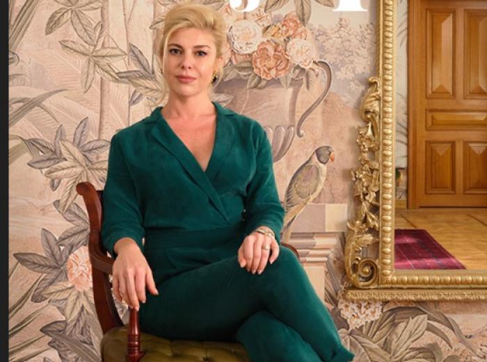 Gözde Kansu poartă o salopetă verde închis și își ține picioarele încrucișate