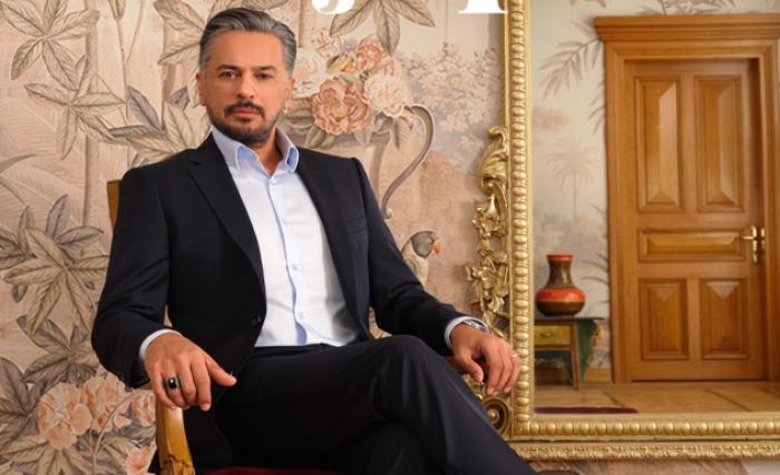 Emre Altuğ poartă o cămașă albă și costum negru și stă pe scaun