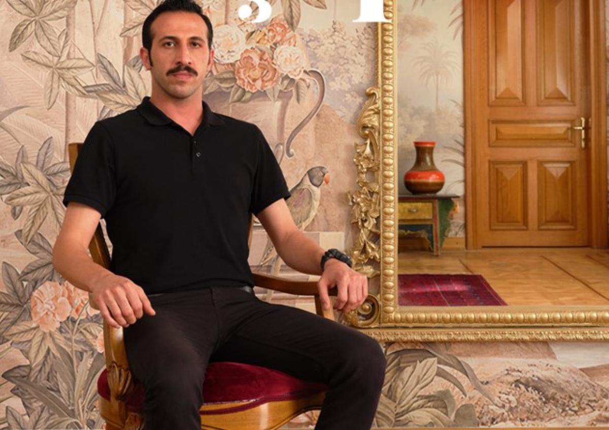 Ersin Arıcı poartă pantaloni negri lungi și un tricou cu mânecă scurtă negru, stă pe scaun și are mustață neagră și părul negru