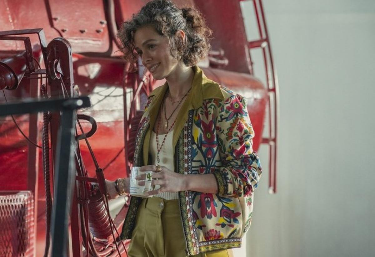 Özge Özpirinççi poartă pantaloni verde muștar, are părul scurt și ține în mâna stângă o cană