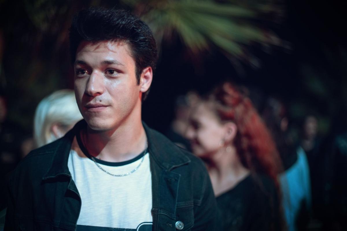 Kubilay Aka poartă un tricou alb și o bluză închisă la culoare