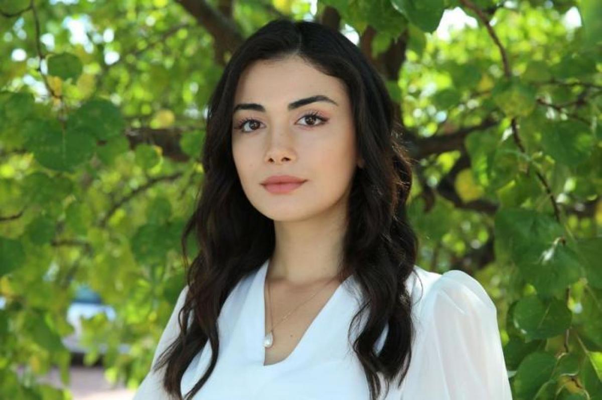 Özge Yağız poartă o rochie albă cu mâneci lungi și are părul lung, brunet și desprins