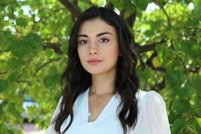 Özge Yağız poartă o rochie albă cu mâneci lungi și are părul lung, brunet și desprins