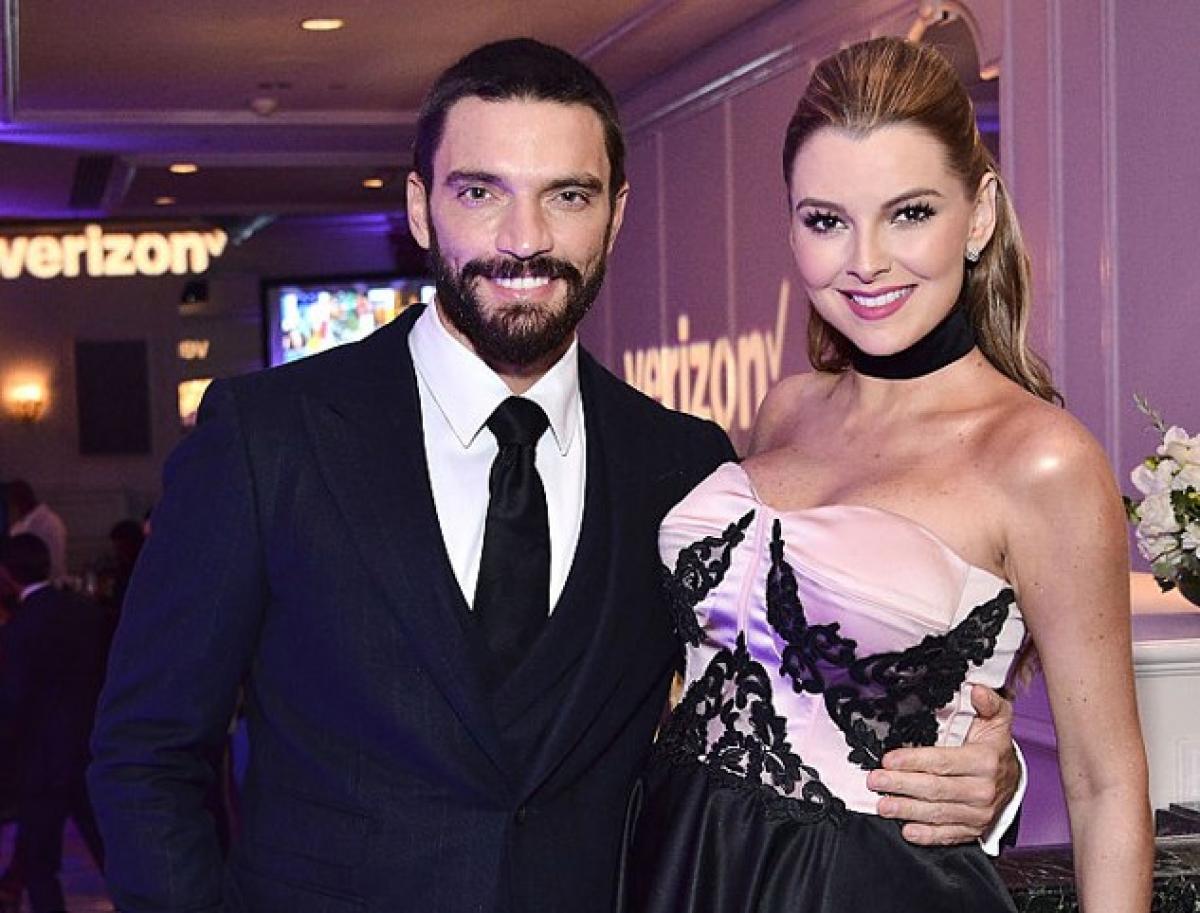 Julian Gil poartă un costum negru, o cămașă albă și cravată neagră și stă alături de Marjorie de Sousa care zâmbește